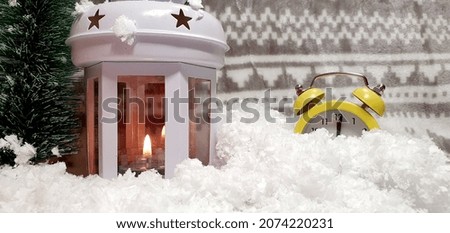Yellow alarm clock in the snow. New Year has come. The hands on the clock point to twelve.
Christmas and New Year decorations, fir tree, lamp with a burning candle, festive winter background.