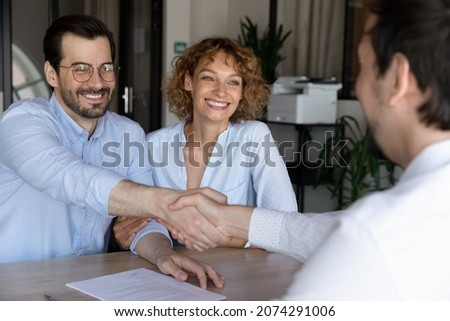 Joyful beautiful young family couple shaking hands with hispanic lawyer or broker, feeling thankful for professional consultation. Happy clients making agreement with realtor at office meeting.