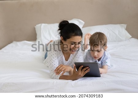 Happy nanny and kid watching movie, media content on tablet together, lying, resting in bed, looking at digital gadget screen, laughing. Mom teaching kid to read, using online learning app