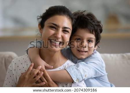 Cheerful cute Indian preschooler kid embracing happy mother on couch, looking at camera, smiling. Young mom and kid hugging with love, affection, tenderness. Head shot home portrait Royalty-Free Stock Photo #2074309186