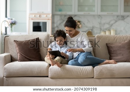 Happy millennial Indian mom gen Z son boy reading book, playing videogame on tablet computer, using online app, service, chatting. Mother and kid shopping, booking on internet at home