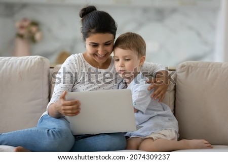 Curious kid and happy nanny holding laptop on sofa, watching online media content, playing games on internet together. Mom teaching son, using learning app on computer, reading book