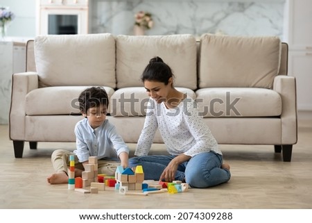Happy young Indian mom helping preschool son to build toy towers from wooden construction blocks, playing with kid on heating floor at home. Babysitter watching child. Motherhood, daycare concept Royalty-Free Stock Photo #2074309288