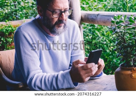 Adult bearded man with blue sweater look at the phone and text message. Modern people with mobile online cellular technology sitting outside home with garden in background