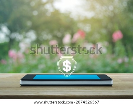 Dollar with shield flat icon on modern smart mobile phone screen on wooden table over blur pink flower and tree in park, Business money insurance and protection online concept