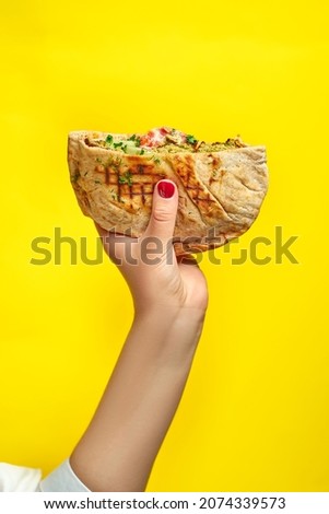 Hand holds pita with chicken and vegetables on a yellow background. Shawarma with chicken