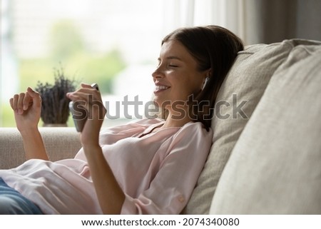 Side view relaxed carefree millennial woman holding cellphone in hands, listening music from mobile library application in wired earphones, enjoying leisure weekend peaceful pastime alone at home.