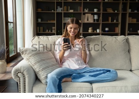 Happy relaxed beautiful young woman in wired earphones looking at cellphone screen, holding video call conversation, communicating distantly or listening music in app, sitting on cozy couch at home. Royalty-Free Stock Photo #2074340227
