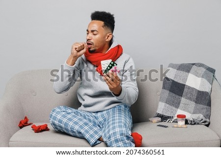 Young ailing african man in grey sweater scarf sit on sofa hold medication tablets pills coughs isolated on plain gray background studio portrait. Healthy lifestyle ill sick disease treatment concept
