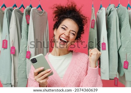 Young happy female costumer woman 20s wear sweater stand near clothes rack with tag sale in store showroom hold in hand use mobile cell phone dow inner gesture isolated on plain pink background studio