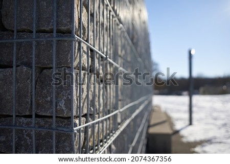 Gabion fence made of steel bars filled with gray granite stones. Empty park bench and a pillar in the snow. Blue sky. Side view. Winter in Germany.