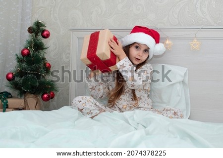 Christmas morning, little girl in pajamas in bed rejoices with gifts on the background of a Christmas tree. A happy, smiling child opens a New Year's gift at home. The concept of holidays, Christmas.