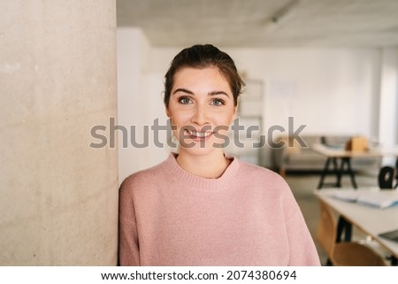 Quiet attentive young woman looking at the camera with an interested or intrigued expression and smile as she leans against a pillar in the office Royalty-Free Stock Photo #2074380694