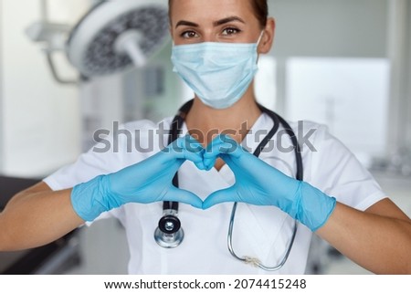 Woman with a medical mask and hands in latex glove shows the symbol of the heart. Doctor for the heart. Love our medical professionals.