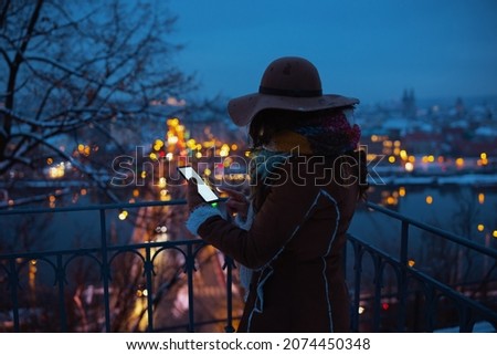 Seen from behind middle aged woman in brown hat and scarf outdoors in the city park in winter in sheepskin coat using smartphone applications at night.