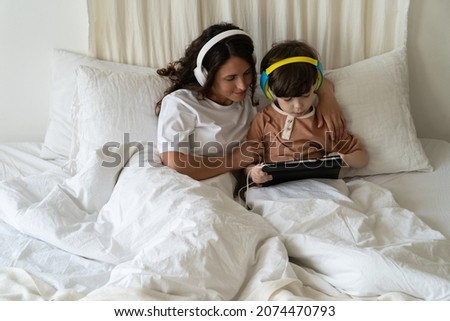 Family morning in bedroom: mom look at little kid son play games on tablet computer or watch video cartoons wearing headphones and listening to music lying with child in bed under blanket on weekend