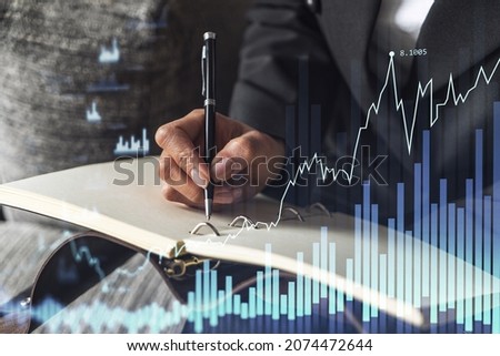 A woman trader in formal wear writing down some ideas to research stock market to proceed right investment solutions. Wealth management concept. Hologram Forex chart over close up shot.