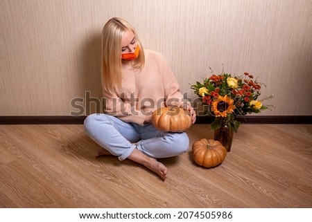 Unhappy young blonde woman wearing medical mask in warm sweater is sitting on the floor and holding a pumpkin in her hands, autumn bouquet with a sunflower nearby.