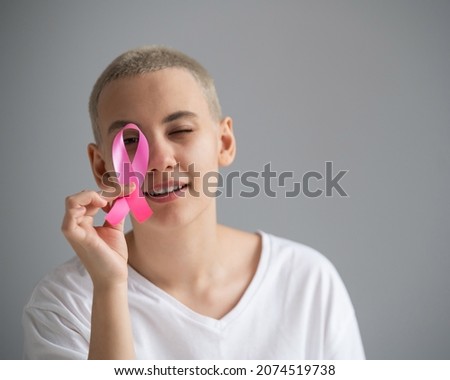 Young woman with short hair wearing a white t-shirt holding a pink ribbon as a symbol of breast cancer on a white background.