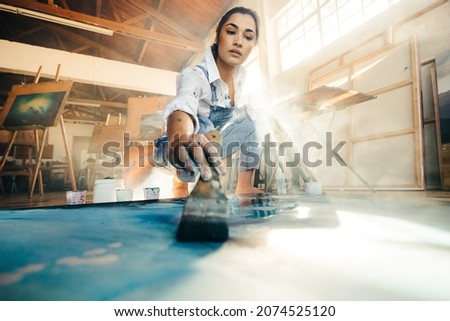 Skilful young artist painting on a canvas with a paintbrush. Female painter making a blue painting for her new art project. Creative young woman working on the floor in her art studio. Royalty-Free Stock Photo #2074525120