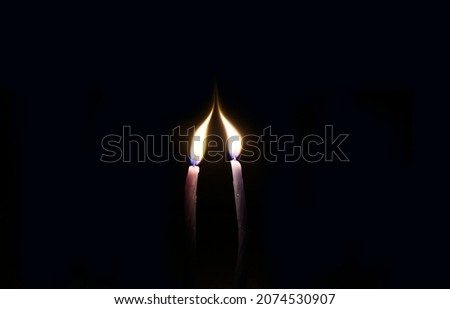 Two thin candles stretch their lights towards each other. The candles are in complete darkness. Black background. 