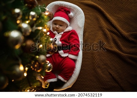 Closeup portrait of newborn baby. Cute Caucasian baby girl 4-5 months old in Santa costume lying on knitted cozy blanket in cocoon near decorated fir tree. Merry Christmas xmas and happy new year 2022