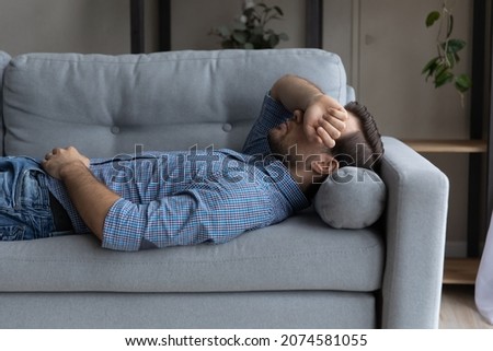 Stressed young man lying on sofa on pillow, covering face with hand, suffering from negative thoughts or insomnia, having lack of energy, sleeping daydreaming napping at home, recreation concept. Royalty-Free Stock Photo #2074581055