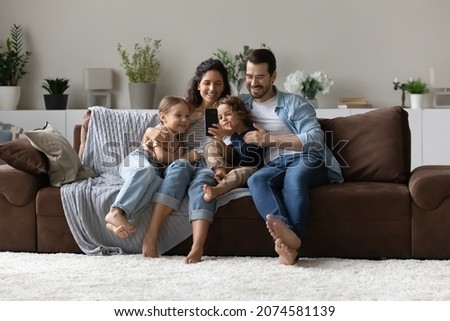 Happy parents with two kids using smartphone at home together, sitting on cozy couch, smiling mother and father with little son and daughter looking at phone screen, taking selfie, having fun Royalty-Free Stock Photo #2074581139