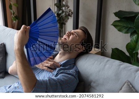 Exhausted young man waving paper fan, breathing fresh cooled air, suffering from high temperature in living room, feeling thirsty overheated indoors without air conditioner system, lying on sofa. Royalty-Free Stock Photo #2074581292