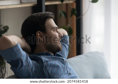 Relaxed happy carefree millennial generation man sitting on cozy couch, breathing fresh air looking in distance daydreaming meditating, visualizing future or planning weekend, enjoying peaceful time. Royalty-Free Stock Photo #2074581418