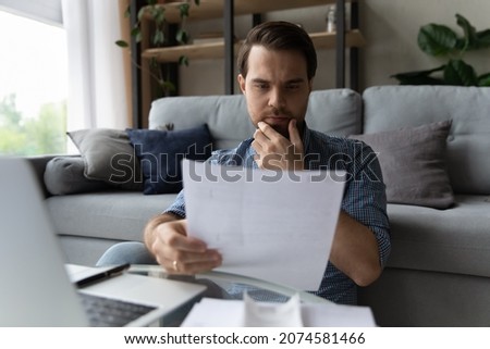 Pensive young businessman reading paper document, considering contract terms. Concentrated male homeowner involved in financial paperwork, analyzing utility bills, thinking of problem solution at home Royalty-Free Stock Photo #2074581466