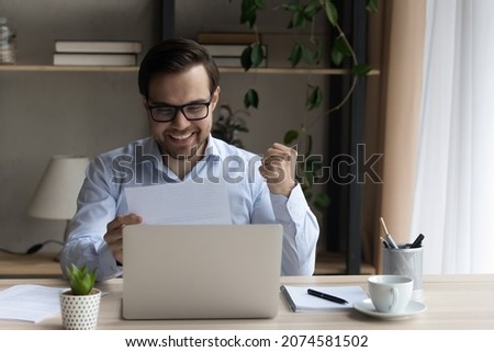 Happy sincere young man in glasses making yes gesture looking at document, reading amazing news in paper correspondence, getting bank loan approval, dream job offer, celebrating signing contract deal.