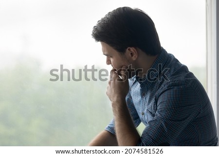 Thoughtful depressed young man touching chin, looking out of window, considering problem solution or feeling stressed of difficult life situation, suffering from negative thoughts alone at home. Royalty-Free Stock Photo #2074581526