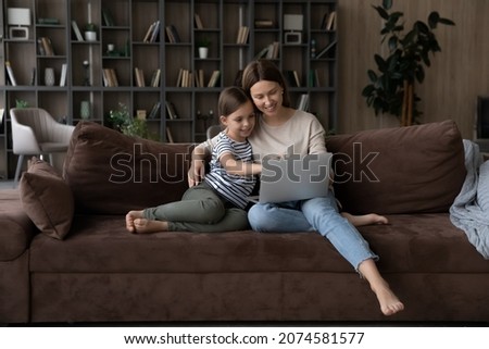 Caring mother with adorable little daughter using laptop at home together, sitting on couch and looking at modern device screen, watching cartoons, video in social networks, having fun online