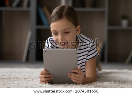 Head shot little girl using tablet, lying on warm floor alone, smiling kid looking at modern device screen, playing game, watching funny video in social network or cartoons, having fun with gadget