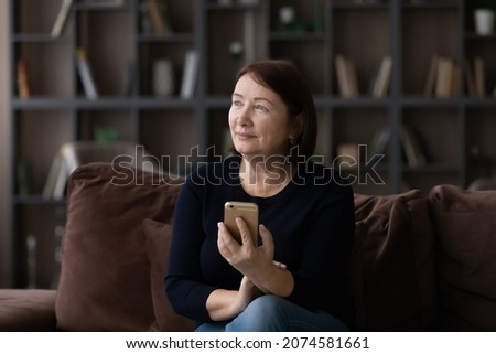 Dreamy mature woman holding modern smartphone in hands, looking to aside, sitting on couch at home, pensive thoughtful senior female visualizing food future, distracted from online communication