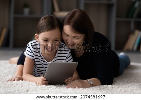 Smiling little girl with mature grandmother using tablet together, lying on warm floor carpet, looking at modern device screen, having fun with gadget at home, watching video, chatting online