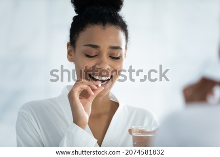 Mirror reflection pretty young african ethnicity woman drinking glass of fresh mineral water, taking pill or daily omega vitamins, dietary supplements for nails hair skincare, support of female health Royalty-Free Stock Photo #2074581832