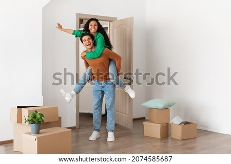Celebrating Moving Day. Happy couple celebrating relocation to their own home, cheerful guy giving lady piggyback ride, excited young millennial family of two having fun standing in new flat Royalty-Free Stock Photo #2074585687