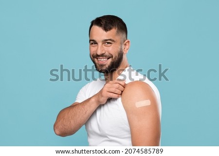 Smiling adult attractive caucasian guy show shoulder with band aid after vaccination, isolated on blue background. Health care, vaccine against flu and covid-19 virus, immunization of population