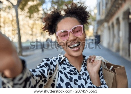 Joyful beautiful woman wears pink sunglasses takes selfie poses at city street with shopping bag feels cheerful and satisfied after making purchasing enjoys free time. People street lifestyle concept