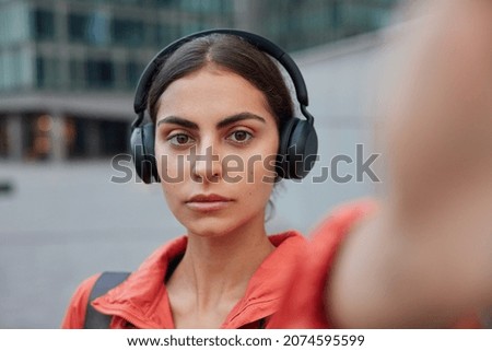 Serious woman stretches arm with uknown device for making selfie listens motivational music for sport in headphones poses against blurred background enjoys favorite audio track strolls during daytime
