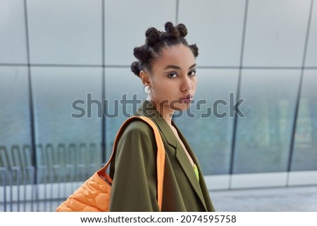 Sideways shot of pretty fashionable woman with hair buns painted blue eyeliners dressed in stylish apparel carries bag has serious expressiong being on way to office poses against blurred background. Royalty-Free Stock Photo #2074595758