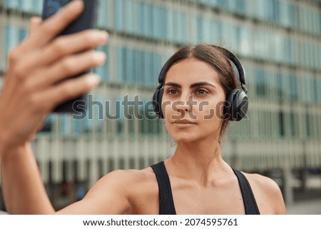 Serious athletic woman uses phone for online call or taking selfie on front camera during fitness workout has confident expression spends free time in urban place poses agains blurred background