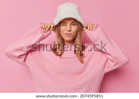 Playful cheerful fashionable girl winks eyes keeps hands on hat wears casual loose jumper poses against pink background dresses for cold weather has fun with friends. People clothes style concept