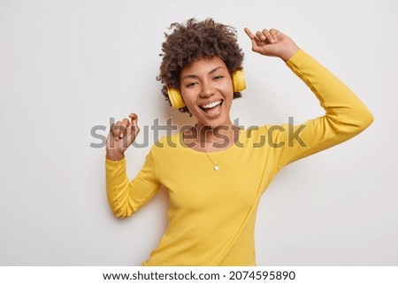 Glad optimistic curly haired woman dances with arms raised up wears casual yellow jumper and wireless headphones has upbeat mood isolated over white background. People emotions and hobby concept