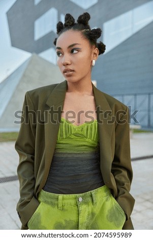 Fashionable woman with bun hairstyle dressed in stylish green clothes keeps hands in pocket looks away pensively poses at urban place during daytime. People street style and lifestyle concept
