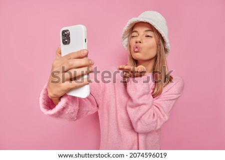 Romantic young woman sends air kiss at cellular camera takes flirty selfie photo winks eye and pouts lips wears hat and jumper isolated over pink background enjoys video chat with boyfriend.