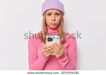 Upset discontent girlfriend waits for call or message from boyfriend feels sad after quarrel and break up wears warm hat pink turtleneck isolated over white background. Negative emotions concept Royalty-Free Stock Photo #2074596100