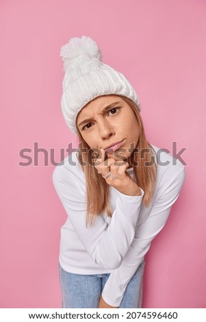 Vertical shot of attentive displeased girl looks seriously at camera wears warm knitted hat and white turtleneck listens unpleasant information keeps finger near lips isolated over pink background